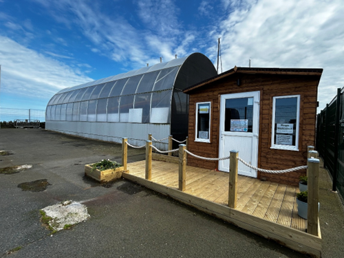 Temporary shelter holding Sir Samuel Kelly. It stands adjacent to a small wooden hut, and has semi-transparent plastic covering, similar to a plastic glasshouse.