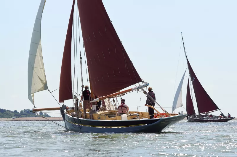 A port stern view of CK437 under full sail on a clear blue sky day. In the background on the right another smack under full sail is shown.