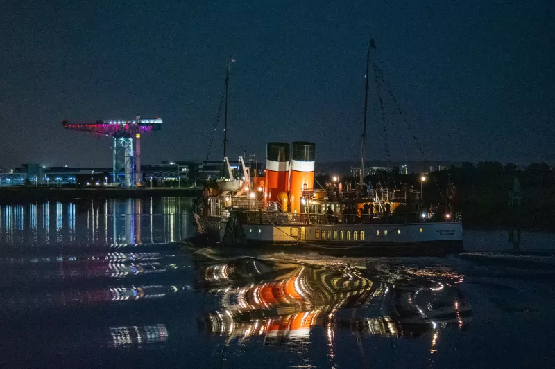 A nighttime shot of PS Waverley, shown from the port stern view, her deck lit up. A tall port crane lit up in light blue, pink, and red is in the background