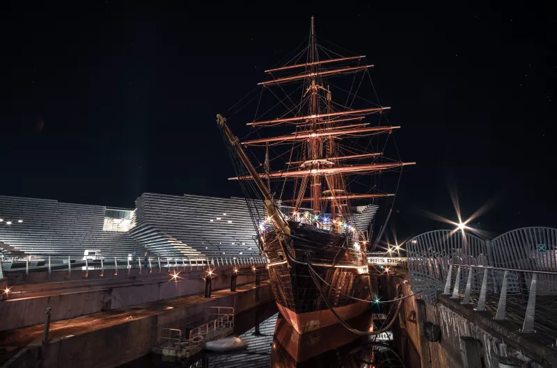 RRS Discovery is shown from the port bow at night, her barren masts lit up from the colourful lighs on her deck. Part of the dry dock, as wel as the V&A building behind the ship, are visible.