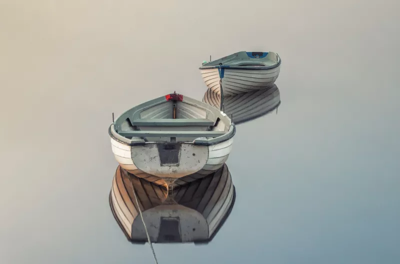 Two small boats with white hulls and duck blue fittings are standing still on a clear and flat water surface. Their hulls are perfectly reflected in the water.