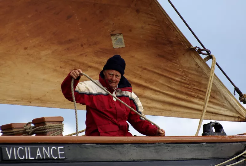 An older man manning the jib sheet line shown against the backdrop of rusty sails